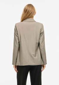 Femme aux cheveux longs blonds portant un blazer taupe et un pantalon noir, debout de dos devant un fond blanc uni.