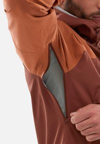 Veste imperméable dans des tons bruns terreux avec un design coloré, dotée d'une poche zippée, d'une texture lisse et d'une doublure respirante.