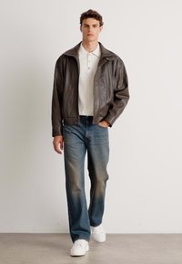 Chaqueta de cuero marrón, camisa polo blanca, jeans azules, zapatillas blancas. La chaqueta cuenta con una cremallera y un cuello amplio, ofreciendo un aspecto casual.