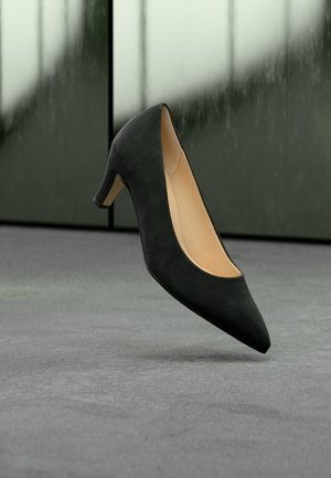 Chaussure femme en daim noir à talon moyen avec bout pointu flottant au-dessus d’un sol gris sur un fond sombre flou.