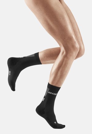 Schwarze Sportsocken mit strukturiertem Design und Logo, die einen engen Sitz um den Knöchel bieten und Fußgewölbeunterstützung haben, in Bewegung an schlanken Beinen dargestellt.