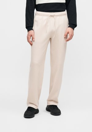 Homme portant un pantalon de jogging beige à cordon et des baskets noires, debout devant un fond clair uni.