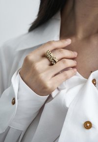 Aukso žiedai su žodžiais "sveiki atvykę" ir "naujas" sukrauti ant rankos, vilkint baltą mygtukinį marškinį su glotnia tekstūra.