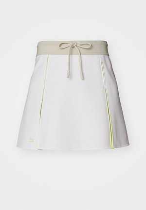 Bílá skorts s béžovým elastickým pasem a šňůrkou. Obsahuje neonově žluté boční akcenty a sklady. Logo Lacoste na dolním levém rohu.