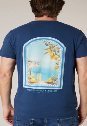 Homme portant un t-shirt bleu marine avec une scène côtière méditerranéenne, des citrons et un verre de limonade imprimés au dos.