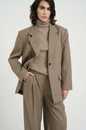 Blazer brun oversize avec un bouton unique, associé à un col roulé beige clair et un pantalon plissé assorti. Texture lisse sur l'ensemble.