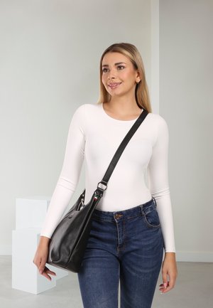 Vrouw in een wit shirt met lange mouwen en blauwe jeans, met een zwarte leren crossbodytas, staand binnenshuis met een neutrale achtergrond.