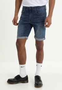 Denim shorts in dark blue with a rolled hem, featuring five pockets and a front button closure, paired with black shoes and white socks.