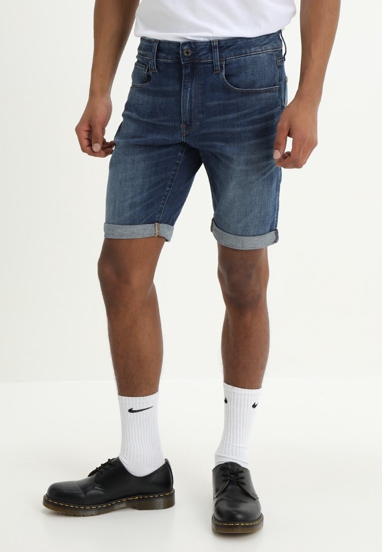 Denim shorts in dark blue with a rolled hem, featuring five pockets and a front button closure, paired with black shoes and white socks.