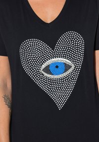 Schwarzes V-Ausschnitt-T-Shirt mit Strassherzdesign. Zentrales Augendetail mit blauen und schwarzen Akzenten, schimmernde Textur mit runden Verzierungen.