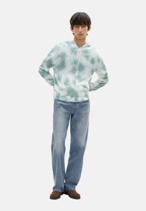 Sudadera con capucha en patrón tie-dye verde claro, cierre frontal con cremallera, dos bolsillos laterales. Combinada con jeans rectos azules y zapatos marrones.