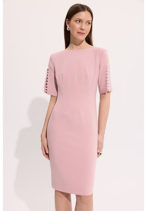 Kobieta w dopasowanej, sięgającej do kolan sukience w pastelowym różu z półdługimi rękawami ozdobionymi rzędami białych guzików, patrząca na bok na jednolitym tle.