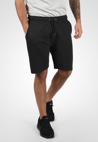 Shorts noirs en coton avec une taille élastique et un cordon de serrage. Texture lisse et poches latérales, associés à des chaussures de sport noires.