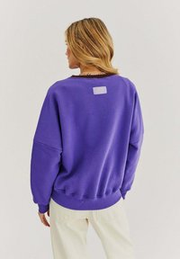 Fialová nadměrná mikina s žebrovanými manžetami a spodním lemem. Na zadní straně se nachází malá nášivka. Měkký bavlněný materiál s hladkou strukturou.