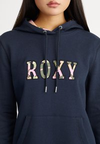 Sweat à capuche bleu marine avec logo "ROXY" sur une patch texturé présentant un motif floral. Cordon de serrage réglable avec embouts métalliques et poche kangourou à l'avant.