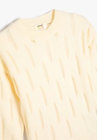 Suéter color crema con un cuello acanalado y recortes en forma de diamante por todo el diseño. Hecho de una suave tela de punto texturizada para mayor comodidad.
