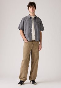Kortarmige geruite shirt in marineblauw en wit, gecombineerd met lichtbruine baggy broeken en zwarte sneakers met witte zolen. Casual uitstraling.