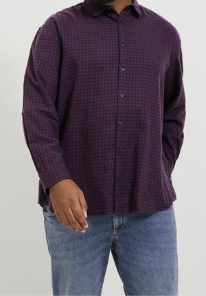 Homme portant une chemise à carreaux rouge foncé et bleu marine à manches longues et un jean bleu clair, debout avec une main légèrement levée.