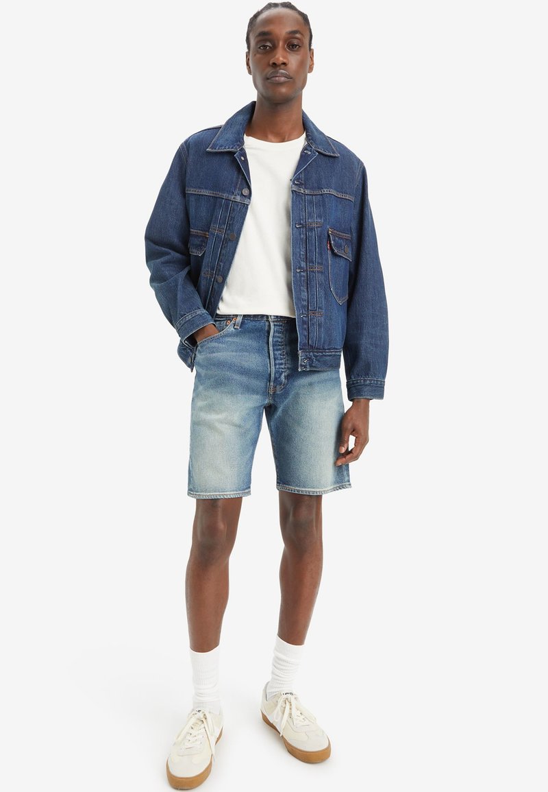 Dunkelblaue Denimjacke, kombiniert mit hellblauen Denimshorts. Darunter ein weißes T-Shirt. Helle Sneakers und weiße Socken runden das Outfit ab.