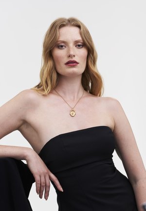 Zlata verižica z okroglim obeskom, ki ima izpopolnjene detajle, nošena prek črnih obleke brez naramnic. Gladek, poliran površinski sloj in topel odtenek.