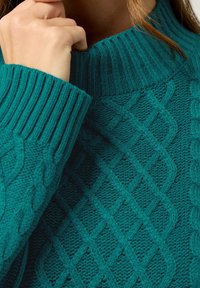Türkiser gestrickter Pullover mit einem gerippten hohen Kragen, der ein strukturiertes Muster aus Zöpfen und Rauten aufweist. Weiche, dicke Garne sorgen für Wärme.