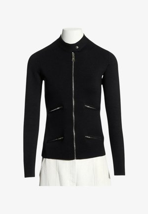 Sort langærmet lynlås-sweater med høj krave og fire vandrette lynlåslommer, vist på en mannequin med hvide bukser.