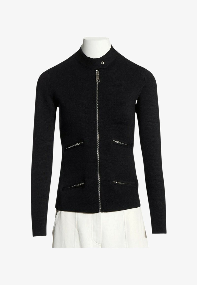 Pull noir zippé à manches longues avec col montant et quatre poches horizontales zippées, montré sur un mannequin portant un pantalon blanc.
