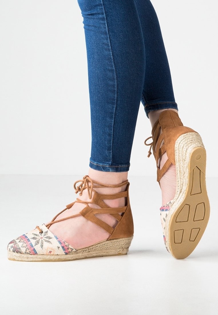 Pieds portant des espadrilles compensées beige avec des lanières à lacets à la cheville et des bouts en tissu à motifs, associées à un jean skinny bleu sur un sol blanc.