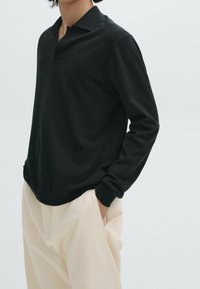 Zwarte polo met lange mouwen, klassieke kraag en ribgebreide manchetten, gecombineerd met lichtbeige broeken. Soepele textuur, casual pasvorm.