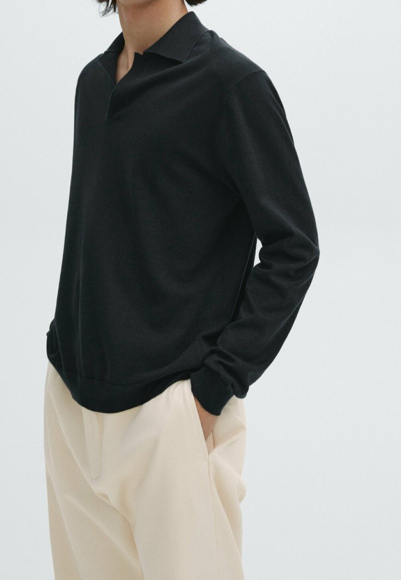 Zwarte polo met lange mouwen, klassieke kraag en ribgebreide manchetten, gecombineerd met lichtbeige broeken. Soepele textuur, casual pasvorm.