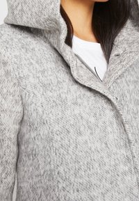 Femme portant un manteau en laine grise texturée avec une grande capuche sur une chemise blanche, montrant la partie supérieure de la poitrine et du cou.