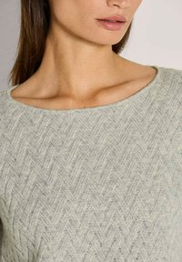 Frau trägt einen hellgrauen, strukturierten Strickpullover mit einem dezenten Zickzackmuster und einem weiten, runden Halsausschnitt.