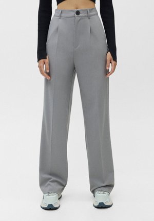 Woman wearing high-waisted, wide-leg gray trousers, black cropped long-sleeve top, and white sneakers, standing against a plain background.