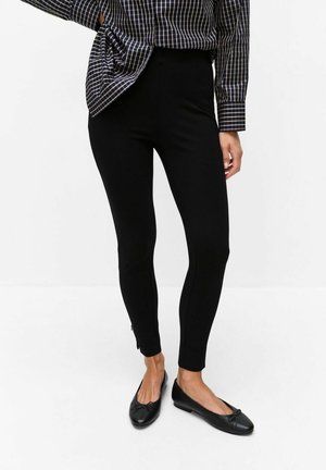 Vrouw draagt zwarte aansluitende enkellange broek, zwarte ballerina's en een geruit overhemd met lange mouwen, met één hand op haar heup.