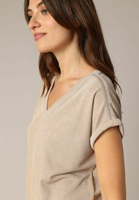 Beige V-hals top met opgerolde korte mouwen. Heeft een zachte textuur en een zilveren accent langs de schoudernaad. Minimalistisch ontwerp.