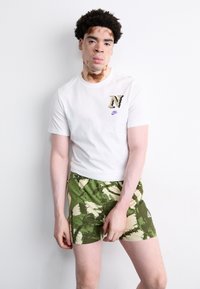 Hvid T-shirt med korte ærmer og et mønstret 'N' samt et lilla Nike-logo. Olivenfarvede shorts med et lysegrønt tropisk bladmønster.