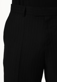 Pantaloni neri a righe sottili con una consistenza liscia, vita aderente e passanti per cintura. Il tessuto ha una leggera lucentezza, enfatizzando il design.