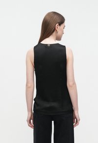 Blusa preta sem mangas com textura suave, apresentando um decote redondo e um pequeno detalhe de logótipo na parte de trás. Usada com jeans pretos.