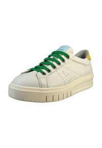 Zapatilla de cuero blanca con cordones verdes, acento amarillo en el talón y suela gruesa con textura. Presenta detalles perforados en el lateral y una etiqueta de color azul claro.