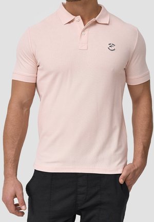 Homme portant un polo à manches courtes rose pâle avec un petit logo sur la poitrine gauche et un pantalon noir, une main dans la poche.