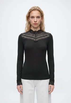Femme aux cheveux blonds portant un haut noir à manches longues avec des détails en dentelle sur la poitrine et les poignets, assorti à un pantalon blanc, debout sur un fond blanc.
