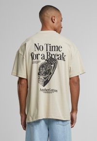Beige bomuld t-shirt med et stort sort grafisk motiv af et ur og teksten "No Time for a Break" på ryggen. Afslappet pasform med korte ærmer.