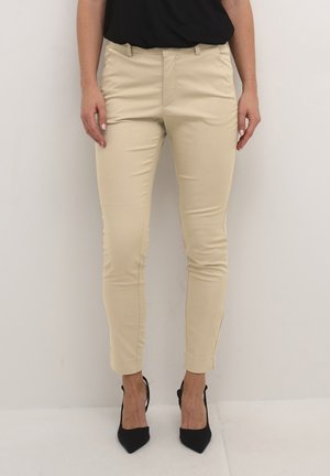 Pantalon fitted beige avec une texture lisse, design à jambe fuselée et petites fentes latérales à l'ourlet, associé à des chaussures à talons hauts noires.