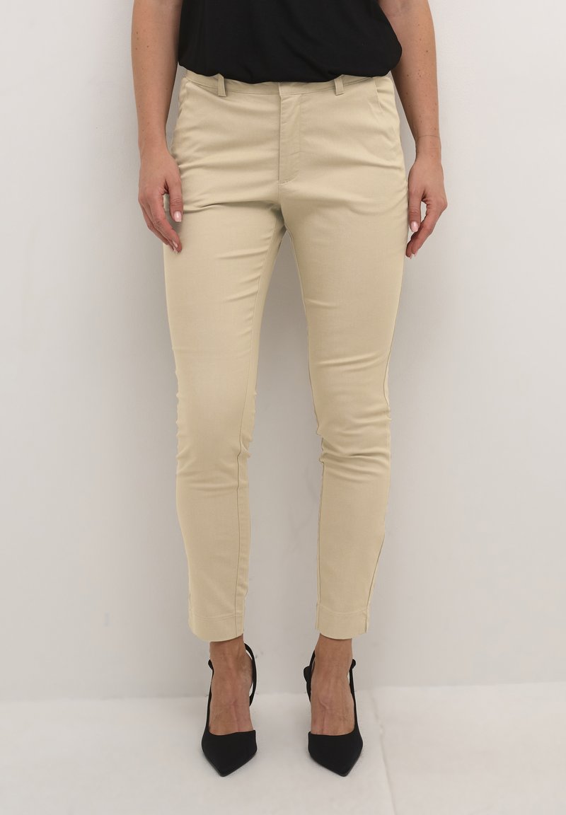 Beige fitted trousers with a smooth texture, tapered leg design, and small side slits at the hem, paired with black high-heeled shoes.