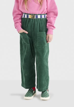 Pantalones de pana verdes con piernas anchas, rayas texturizadas, dos bolsillos laterales, combinados con un suéter rosa y zapatos con lunares, adornados con un cinturón a rayas.