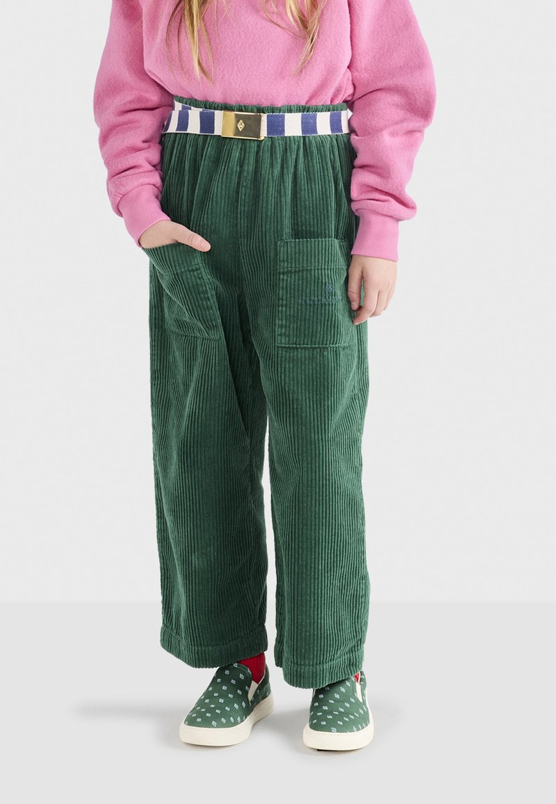 Grüne Cordhosen mit weitem Bein, strukturierten Streifen, zwei Seitentaschen, kombiniert mit einem pinken Pullover und gepunkteten Schuhen, ergänzt durch einen gestreiften Gürtel.