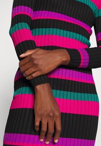 Personne en robe côtelée avec des rayures horizontales pailletées noires, roses, violettes et vertes, les bras croisés à la taille.