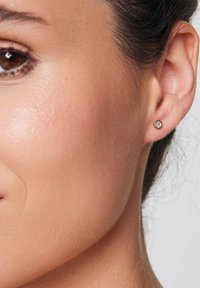 Złote heksagonalne kolczyki wkręty z małym, przezroczystym kamieniem w centrum, o polerowanej powierzchni i subtelnym geometrycznym wzorze.