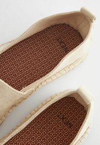 Beige canvas slip-on shoes with rounded toe, textured insole featuring a geometric pattern, and contrasting stitching around the sole.
