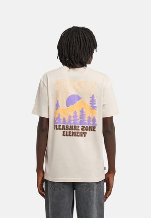 Person mit Dreadlocks, die ein beigefarbenes T-Shirt mit einem lila-orangenen Bergdesign mit Bäumen und dem Text "Pleasure Zone Element" auf der Rückseite trägt.